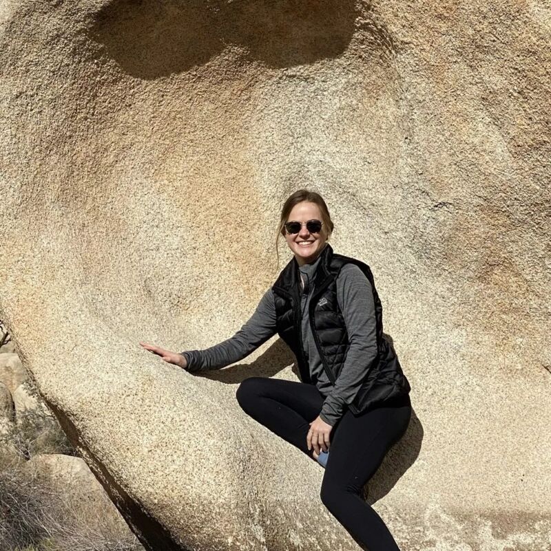 Here is a description of the image: A woman wearing sunglasses, a black vest, and leggings is posing next to a large rock. She is smiling and appears to be enjoying the outdoors. The background suggests a desert environment with sparse vegetation.